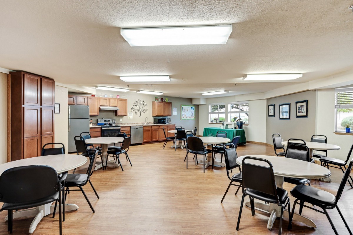 A community room with a kitchen, featuring tables and chairs arranged for social gatherings.