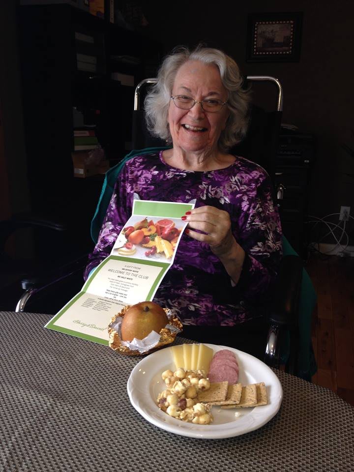 An elderly woman in a wheelchair smiles while holding a card, with a plate of food in front of her.