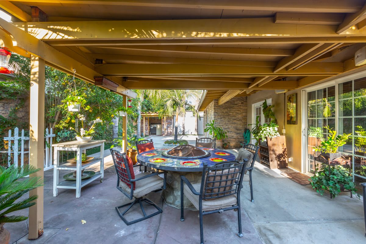 A cozy outdoor patio area with a table and chairs set under a covered pergola, surrounded by lush greenery.
