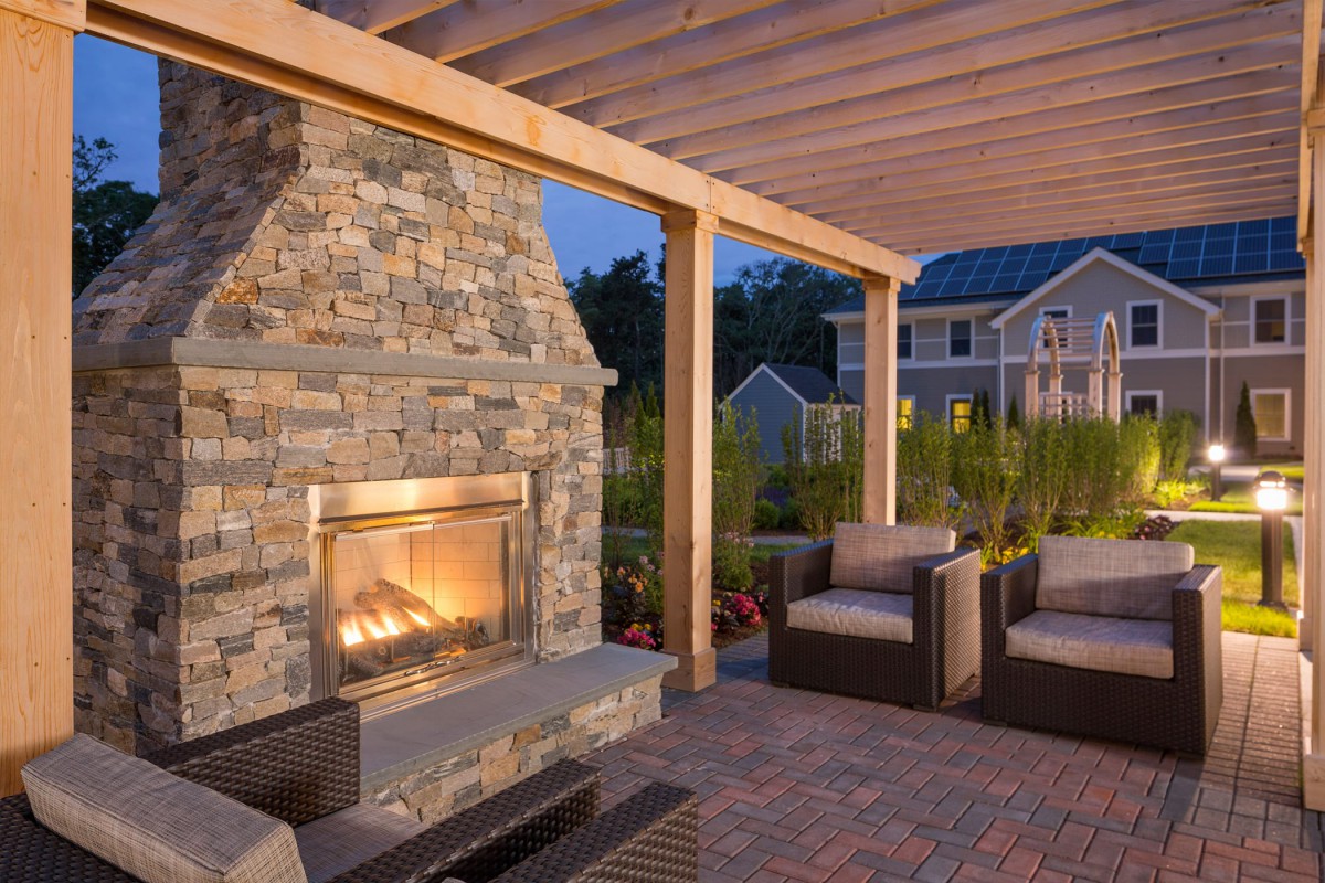 A cozy outdoor patio with a stone fireplace and comfortable seating under a pergola, set for an evening ambiance.