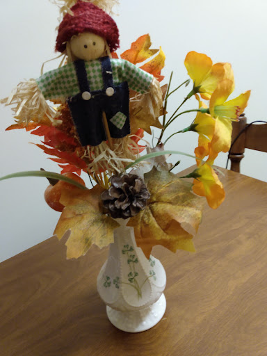 A charming autumn centerpiece featuring a scarecrow and colorful faux flowers in a decorative vase.
