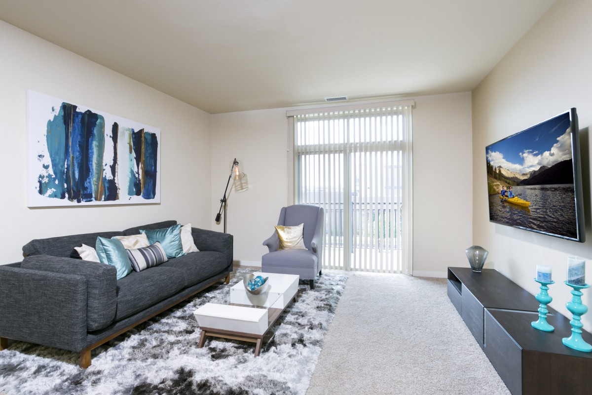 A modern living room with stylish decor, featuring a sofa and armchair, large artwork on the wall, and a floor lamp next to a window with vertical blinds.