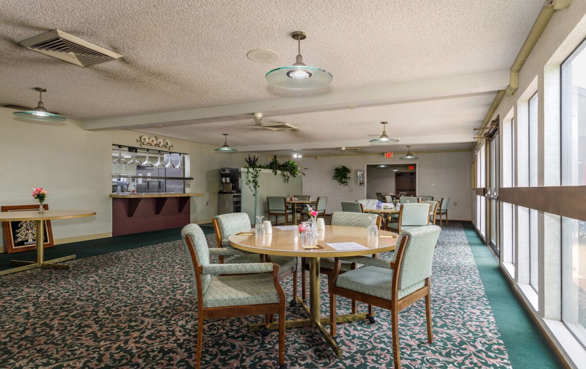 A cozy dining area with tables and chairs arranged on a patterned carpet, complemented by indoor plants and warm lighting.
