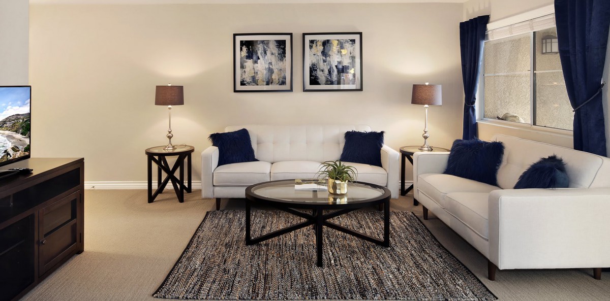 A cozy living room with two white sofas, a round coffee table, and stylish lamps on end tables. The walls feature modern artwork above the seating area.