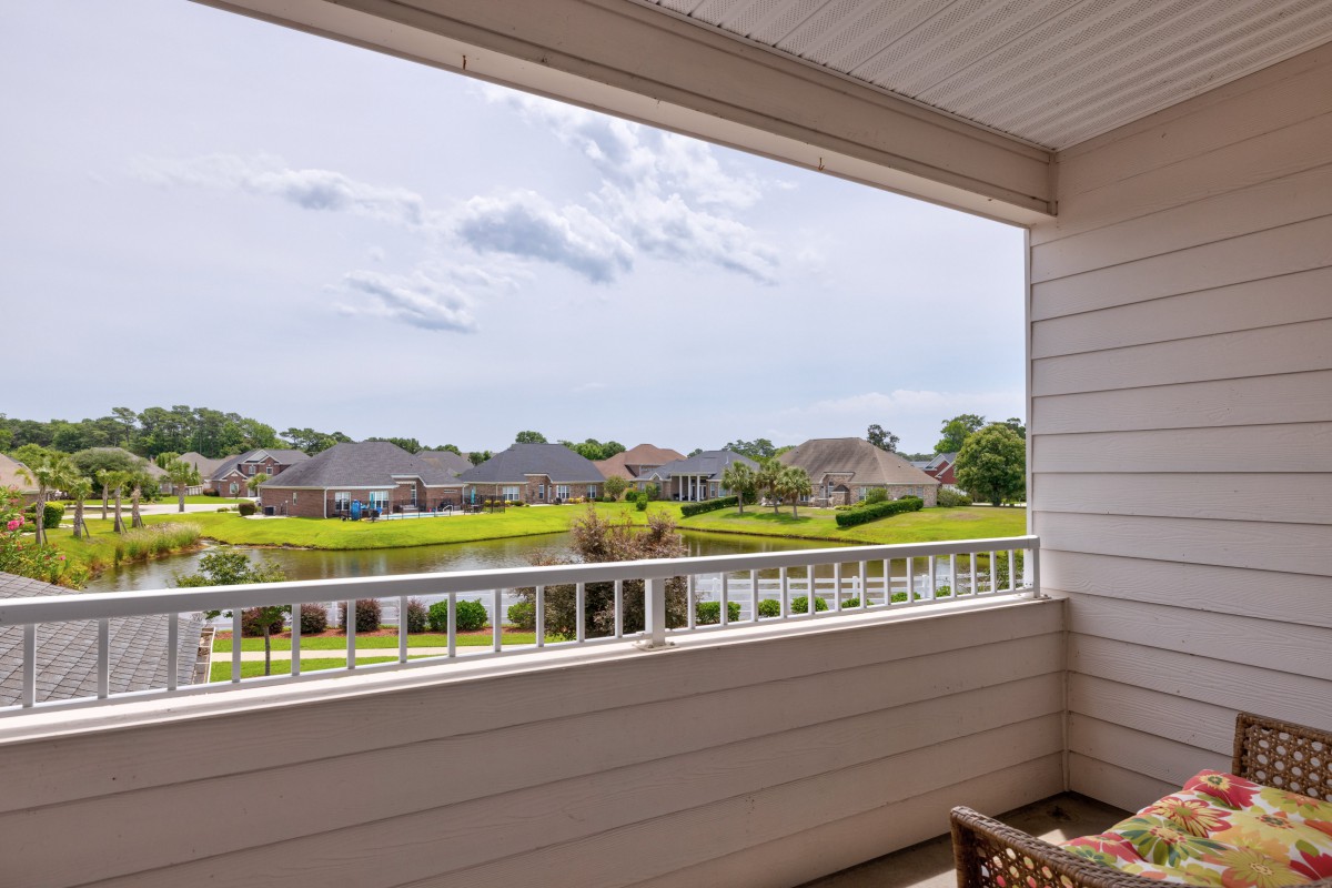 A balcony view overlooking a peaceful residential area with a small lake and houses in the distance under a partly cloudy sky.