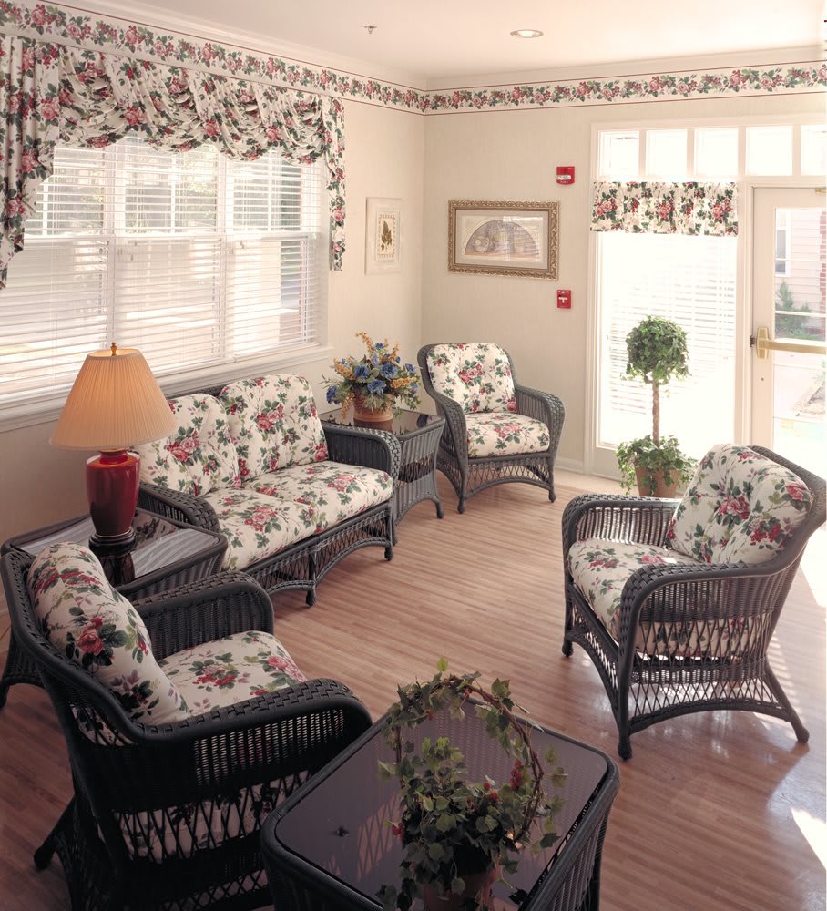 A cozy living room featuring wicker furniture with floral patterned cushions, warmly lit by a lampshade and enhanced by natural light from the windows.