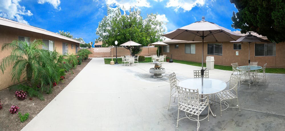 A spacious backyard patio with tables, chairs, and umbrellas under a bright blue sky.
