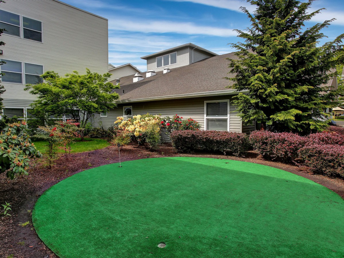 A suburban backyard featuring a well-maintained putting green surrounded by lush landscaping and a house in the background.