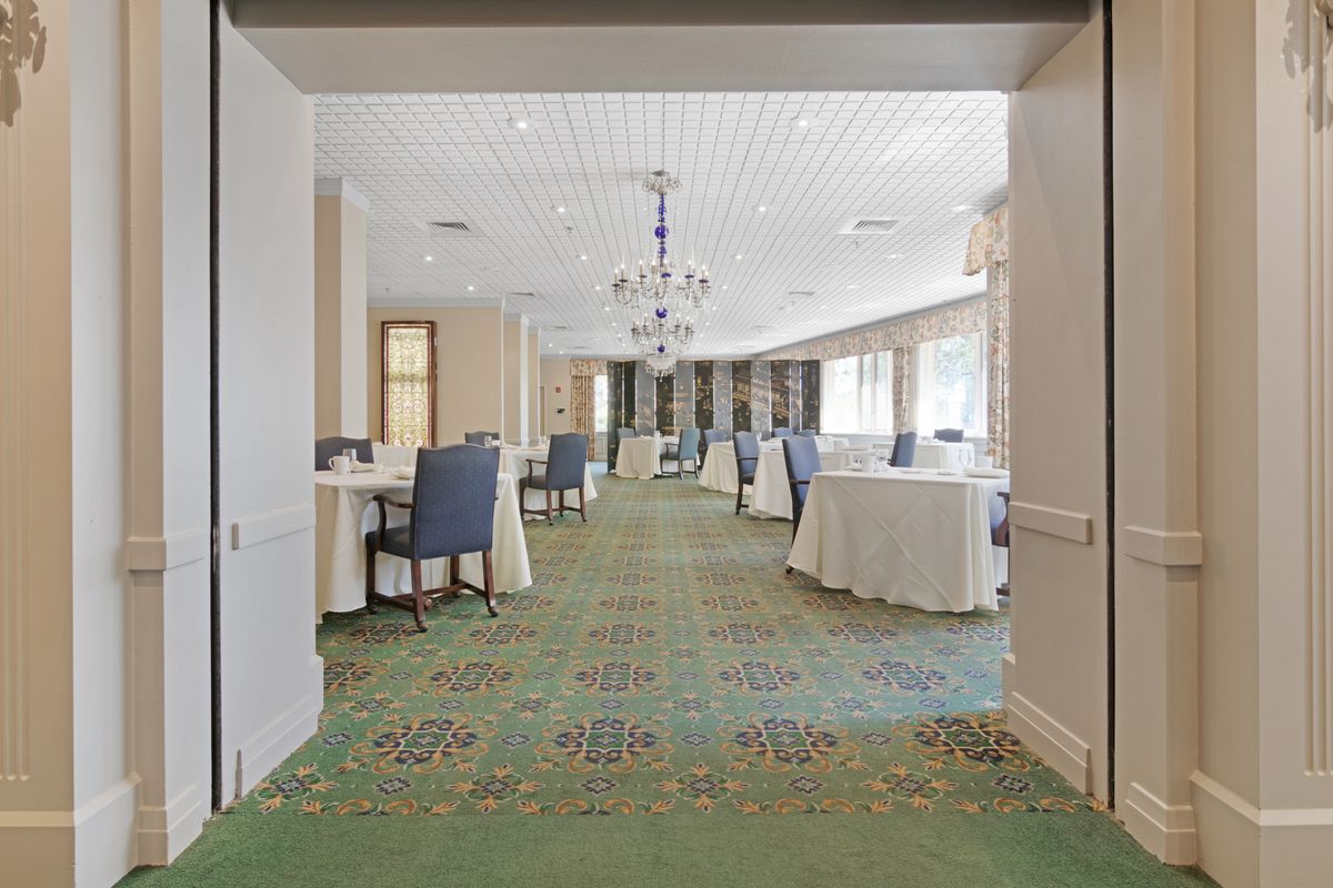 A spacious conference room with round tables and comfortable chairs, illuminated by a large chandelier and natural light.