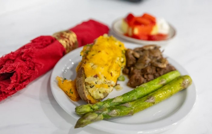 A delicious dinner plate featuring a cheese-topped baked potato, grilled asparagus, and beef steak, accompanied by a side dish with red accents in the background.