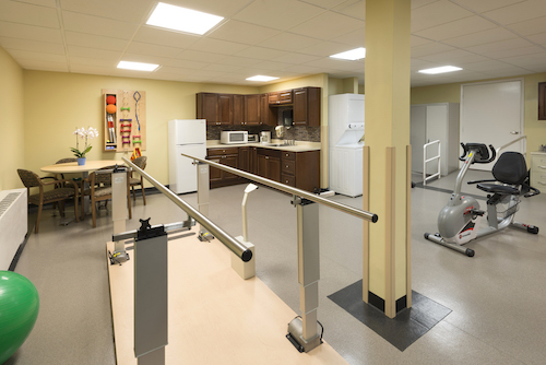 A bright and spacious rehabilitation therapy room featuring parallel bars and a kitchenette.