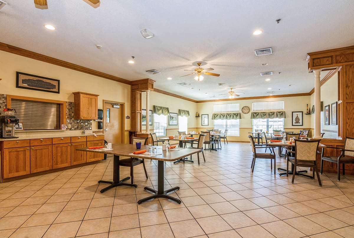 A spacious dining room with several tables and chairs, featuring a kitchen area and large windows allowing natural light.