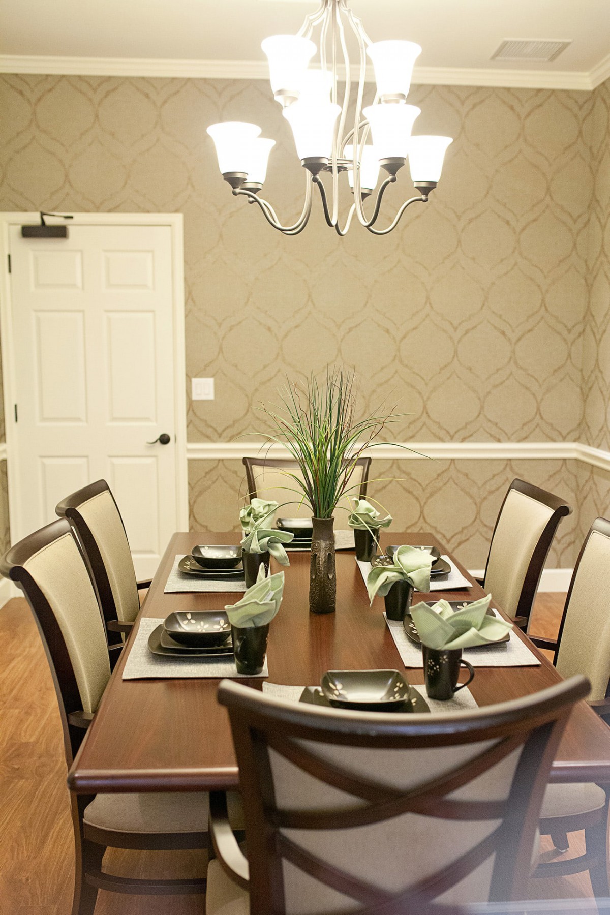 A well-decorated dining room with a wooden table set for six, featuring elegant place settings and central plant decorations under a chandelier.