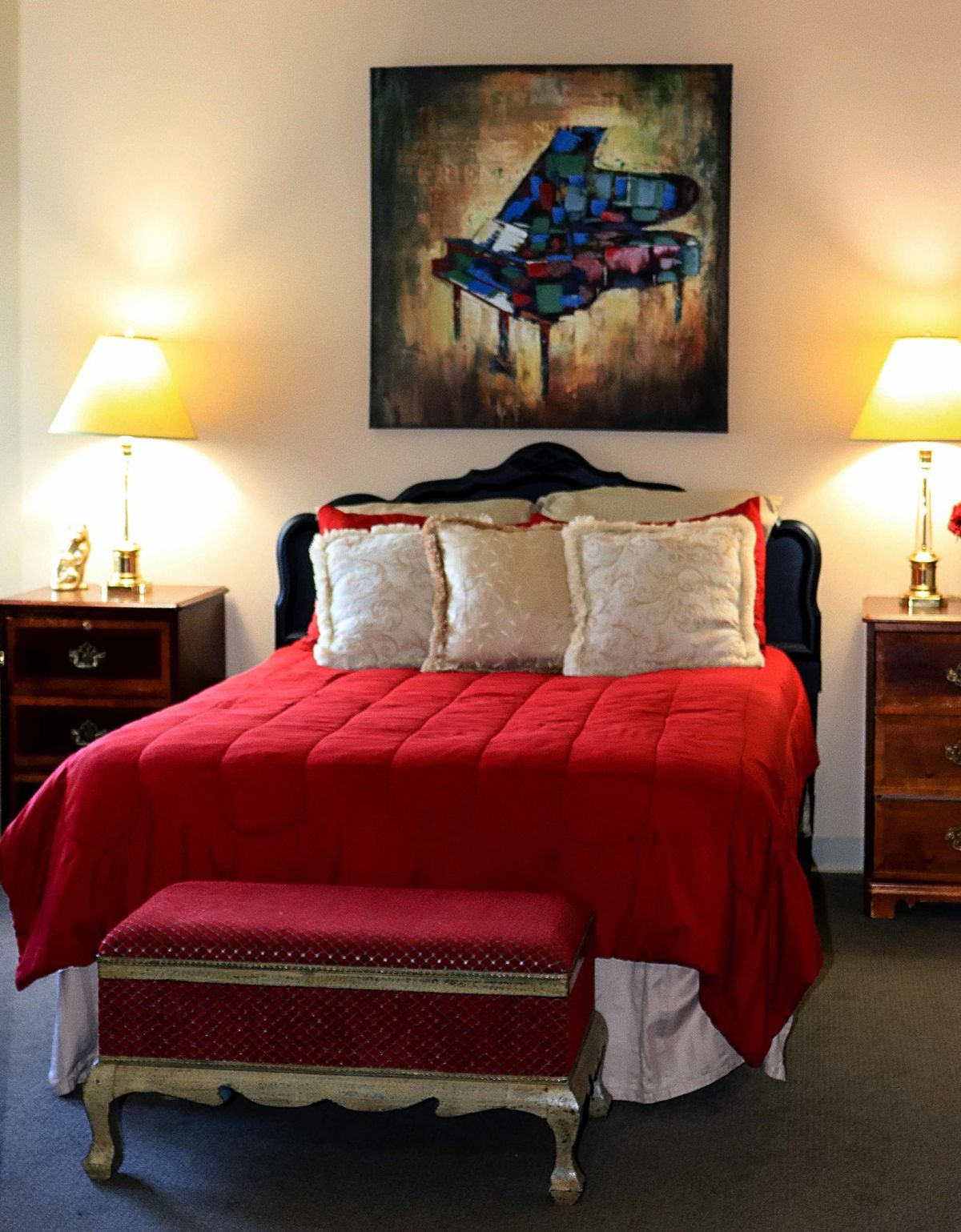 A cozy bedroom with a red bedspread, flanked by nightstands and lamps, and featuring a colorful painting above the headboard.