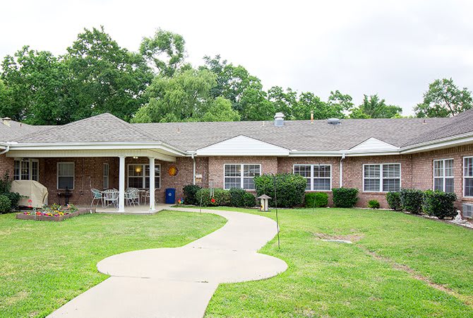 A single-story brick house with a front porch, surrounded by a lawn and trees.