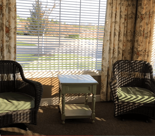 A cozy seating area with two wicker chairs and a small table by a window with blinds.