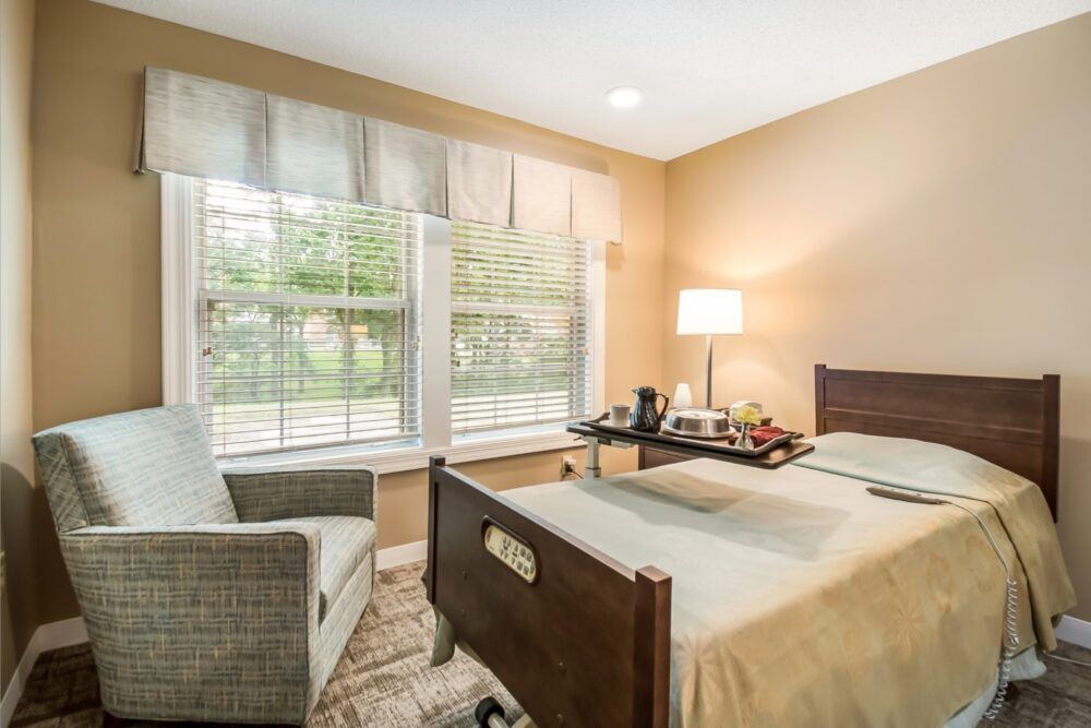A cozy bedroom with a hospital bed, well-lit by a nearby lamp, featuring large windows and comfortable seating.