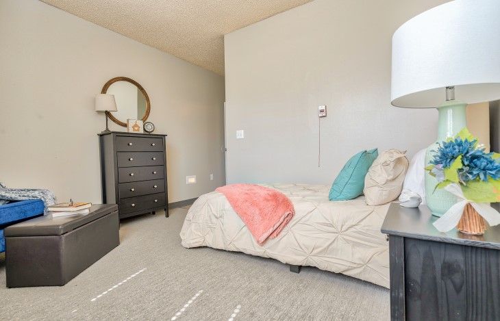 A cozy bedroom with a neatly made bed, featuring colorful pillows and a blanket, alongside a dresser with a round mirror on the wall.