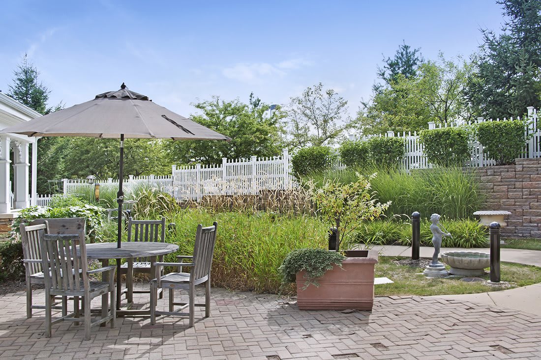 A serene outdoor patio with a table, chairs, and an umbrella surrounded by lush garden greenery.