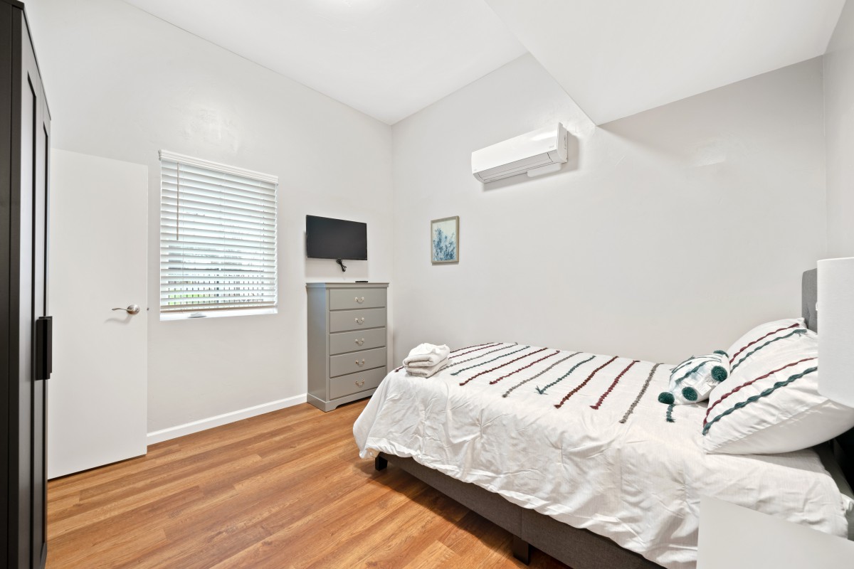 A modern, minimalist bedroom with a bed, dresser, TV, and air conditioning.