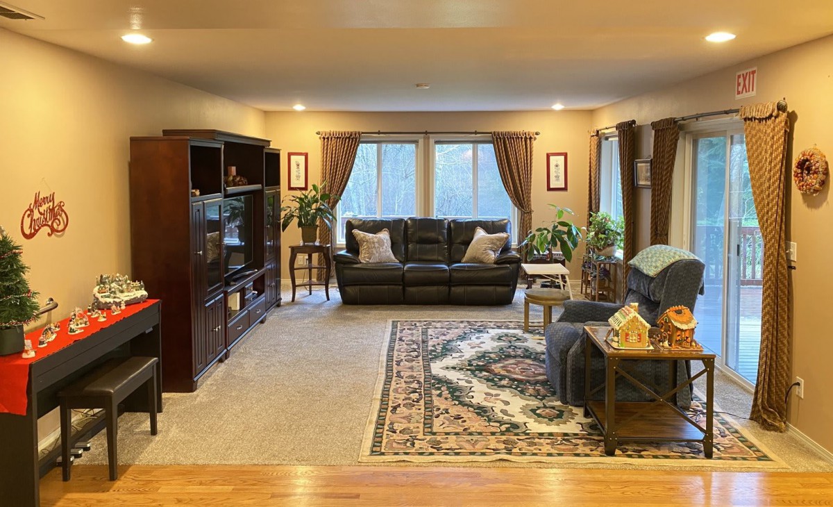 A cozy living room with a sofa, carpet, and bookshelf, featuring large windows and decorative plants.