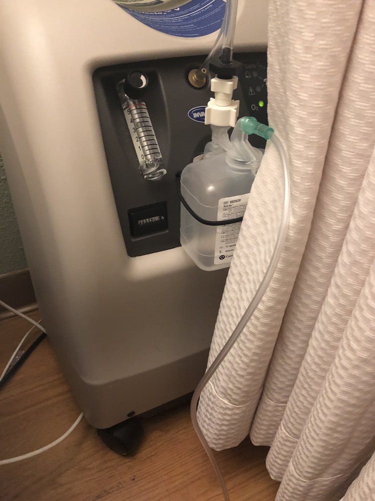 A portable oxygen concentrator with a connected humidifier bottle and tube, placed indoors.