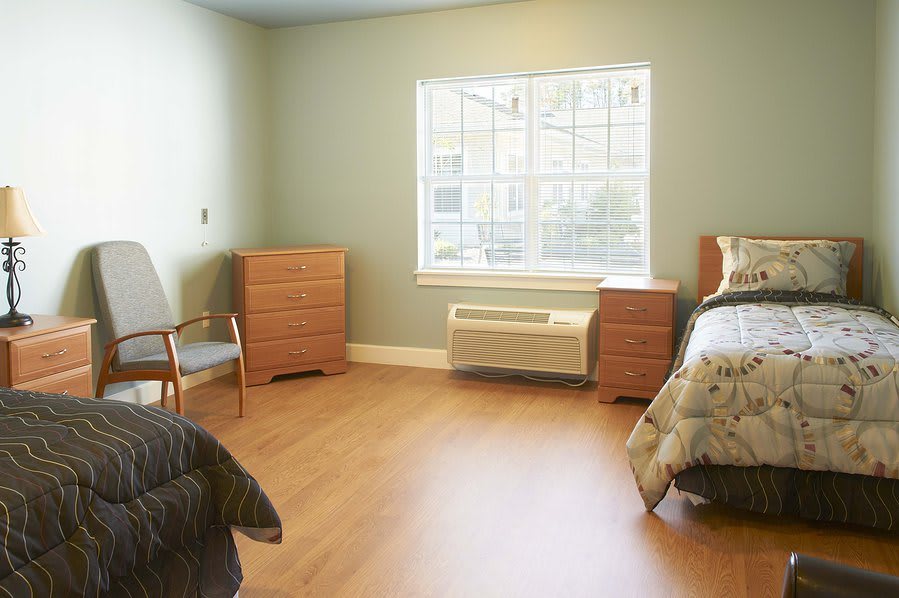 A minimalist bedroom with a bed, two dressers, and a chair, featuring a large window and wooden floors.