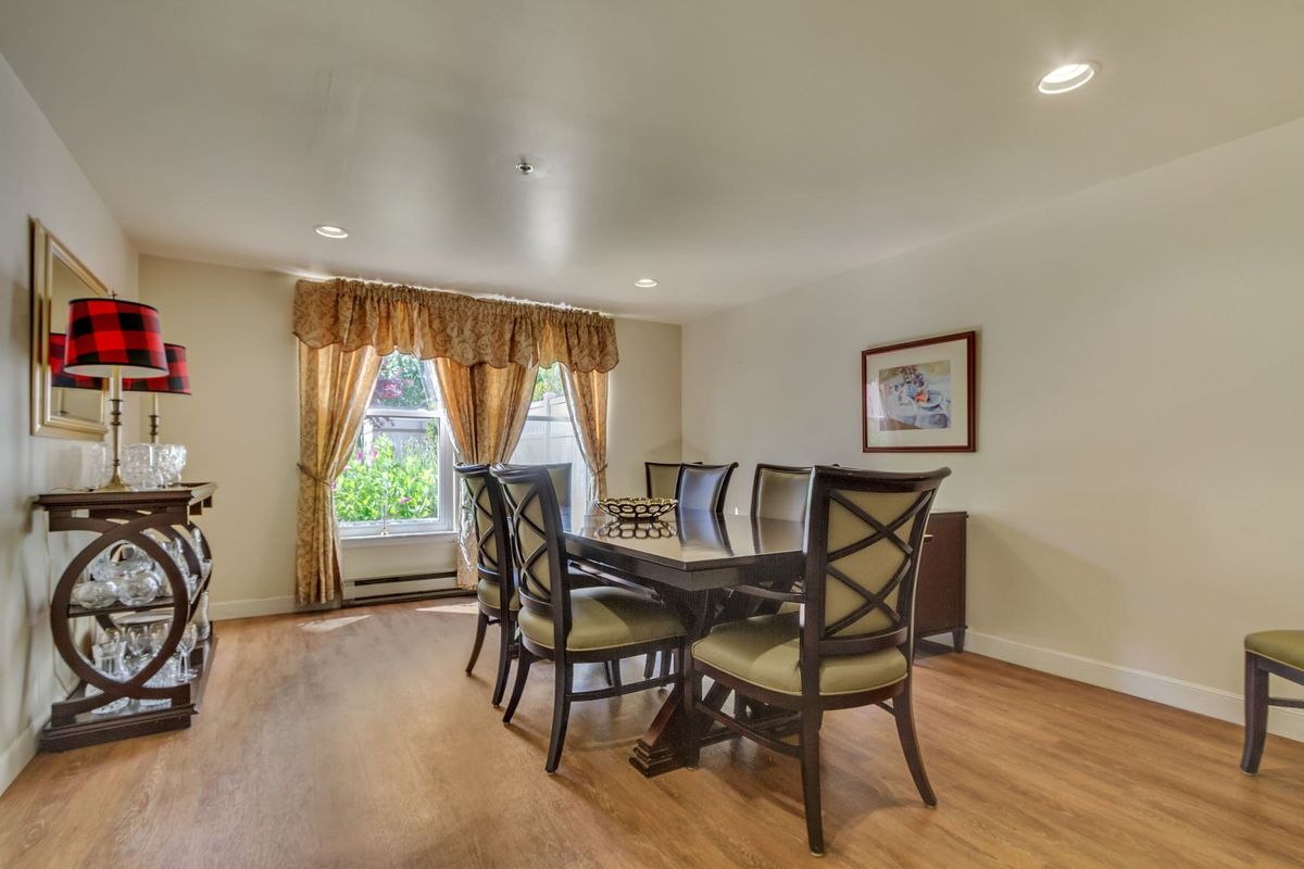 A dining room with a table and chairs, featuring large windows with curtains and a painting on the wall.