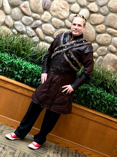 A man in a fantasy-themed costume poses confidently in front of a decorative stone wall and greenery.