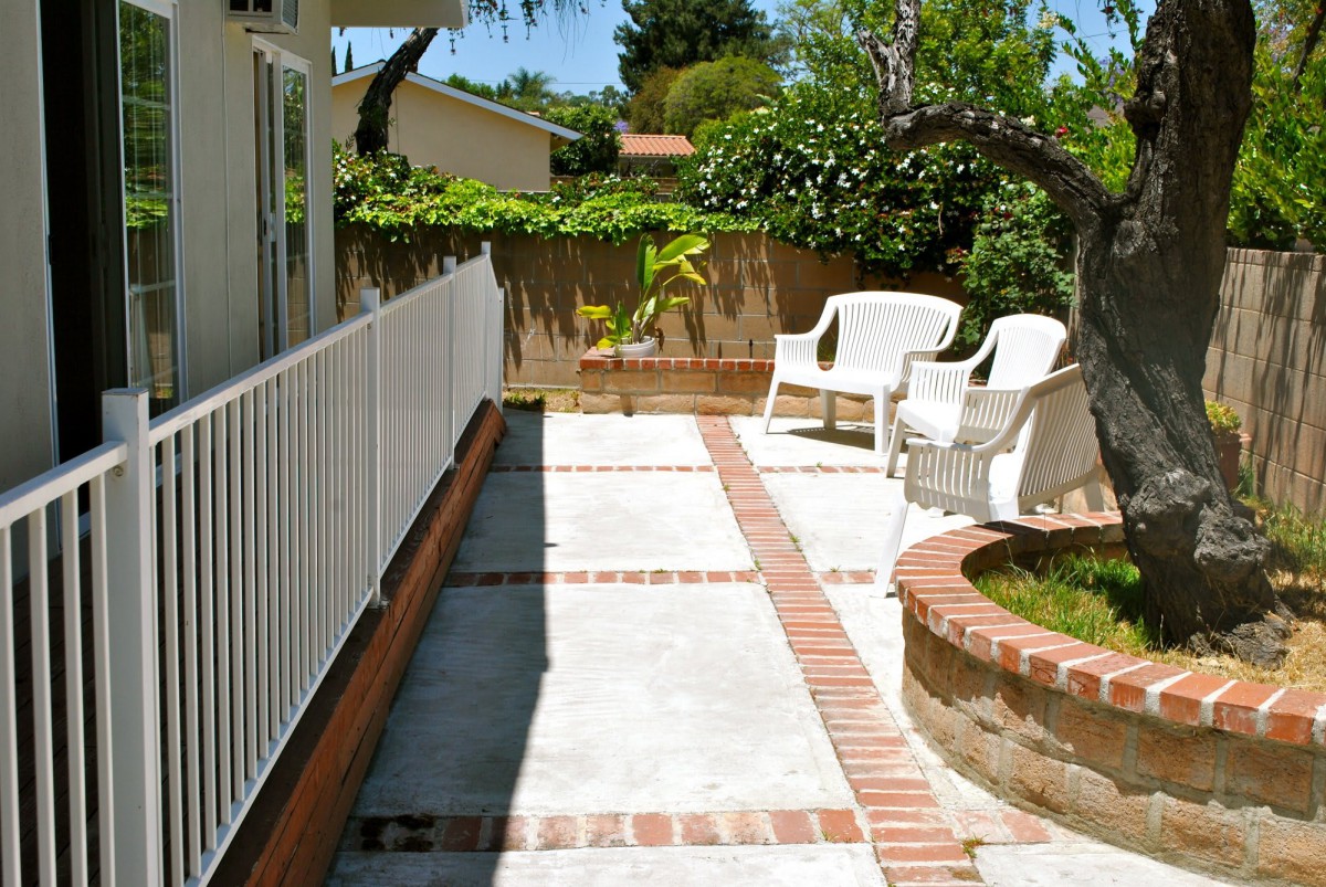 A sunny patio with white chairs, surrounded by a garden and brick pathways.