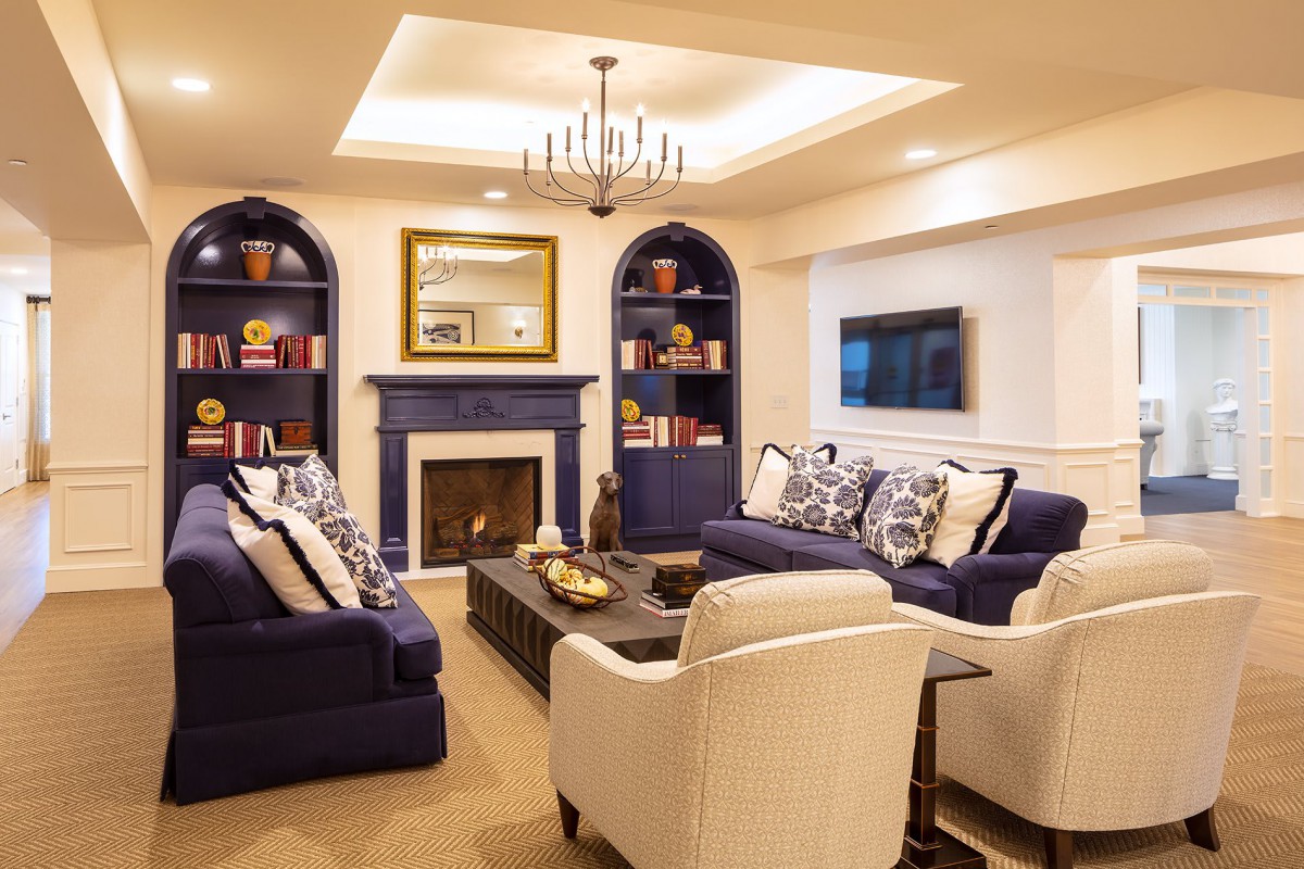 A cozy living room featuring a fireplace, elegant blue and white furniture, built-in bookshelves, and a stylish chandelier.