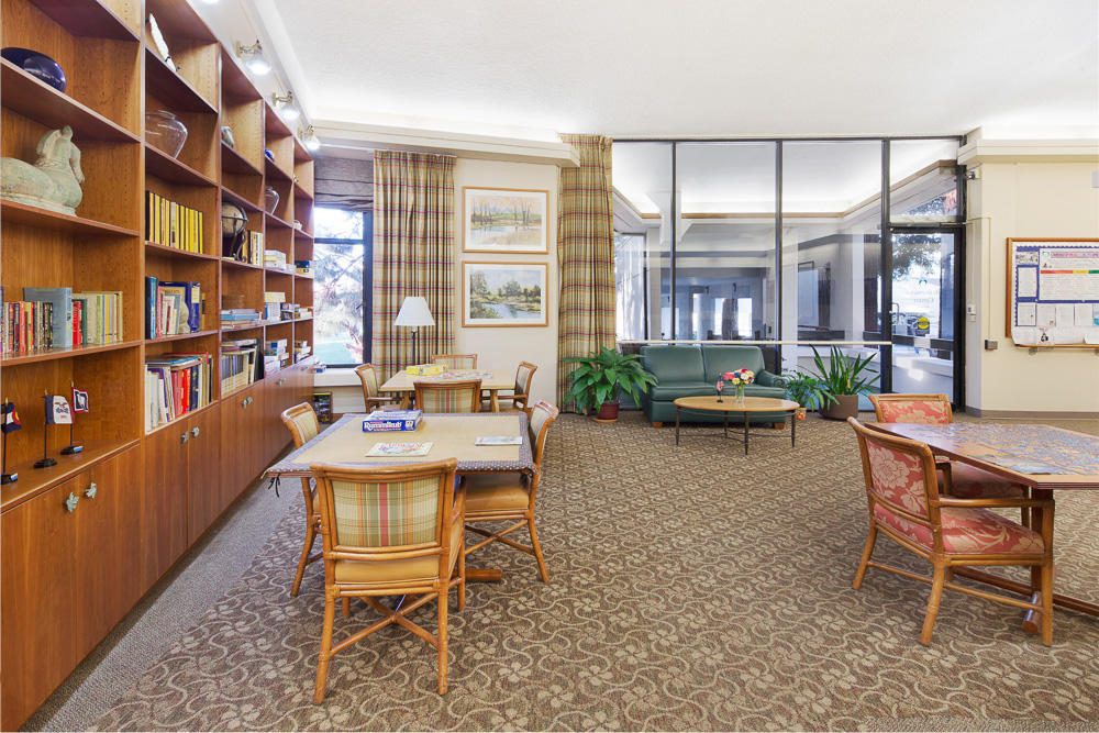 A cozy library interior featuring bookshelves, tables, chairs, and a carpeted floor.