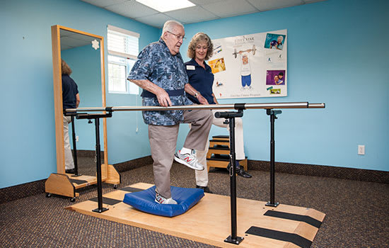 An elderly man practicing balance exercises with the assistance of a healthcare professional in a physical therapy room.