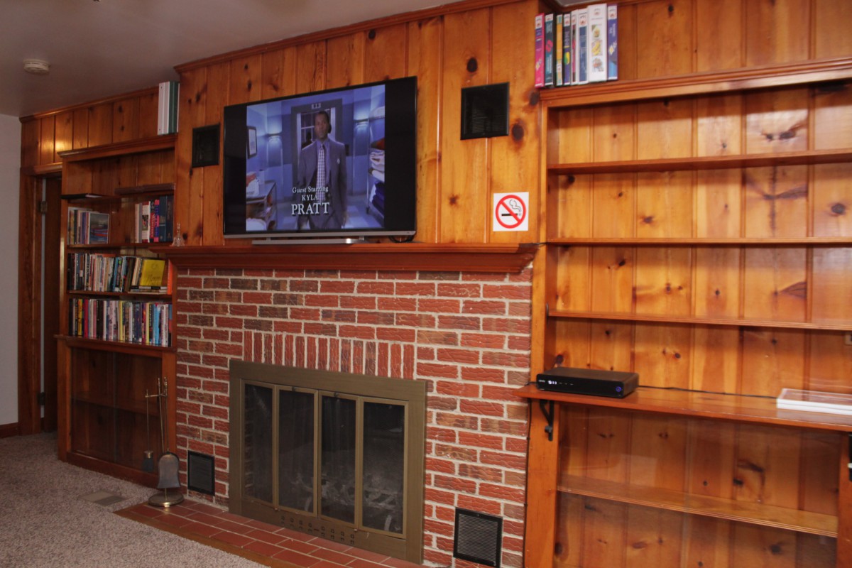 A cozy living room with a brick fireplace, wood-paneled walls, a mounted TV, and built-in shelves.