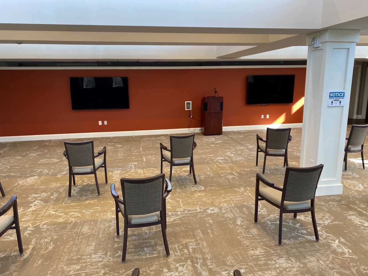 A conference room setup with spaced chairs, a podium, and two mounted television screens.
