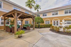 A sunny courtyard with a pergola and outdoor seating, surrounded by palm trees and a two-story building.