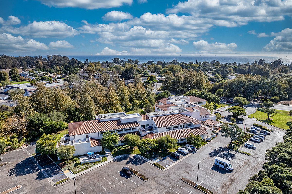 Aerial view of a coastal area with buildings surrounded by trees and a parking lot, under a partly cloudy sky.