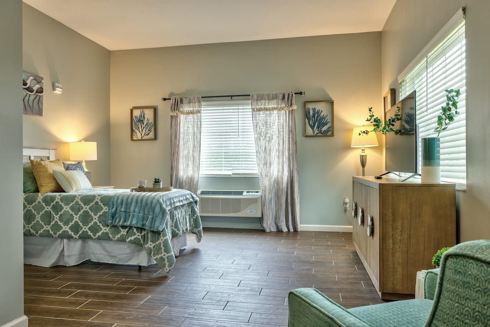 A cozy bedroom with a neatly made bed, framed pictures on the walls, a window with patterned curtains, and soft lighting from lamps.