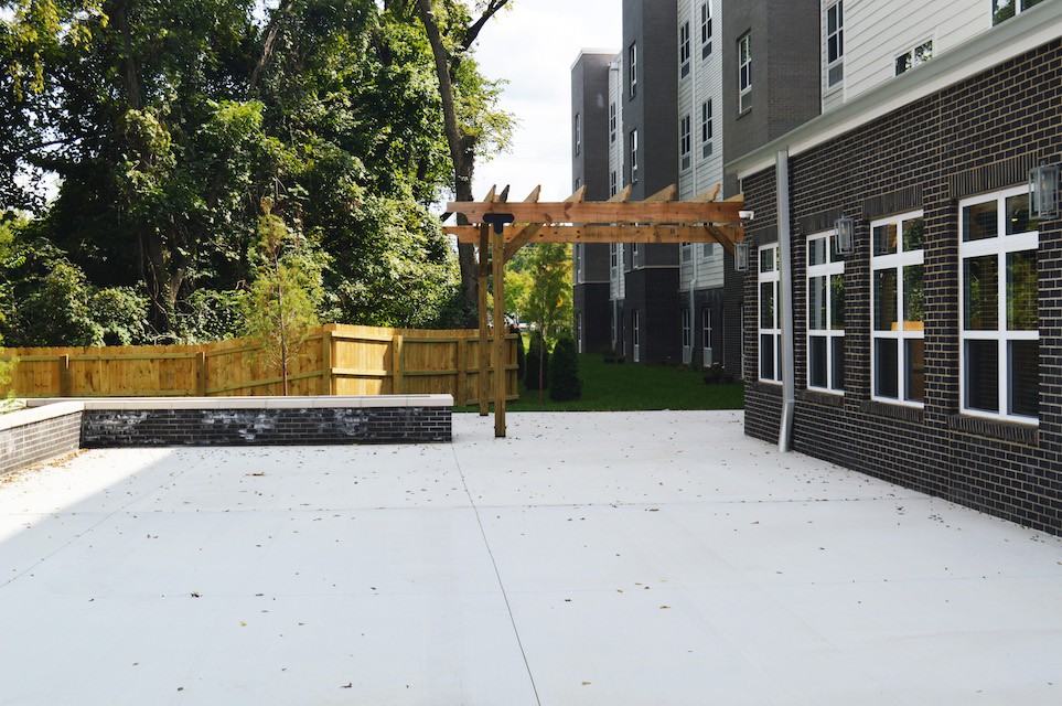 A modern outdoor patio area adjacent to a building, featuring a wooden pergola and surrounded by trees and a fence.