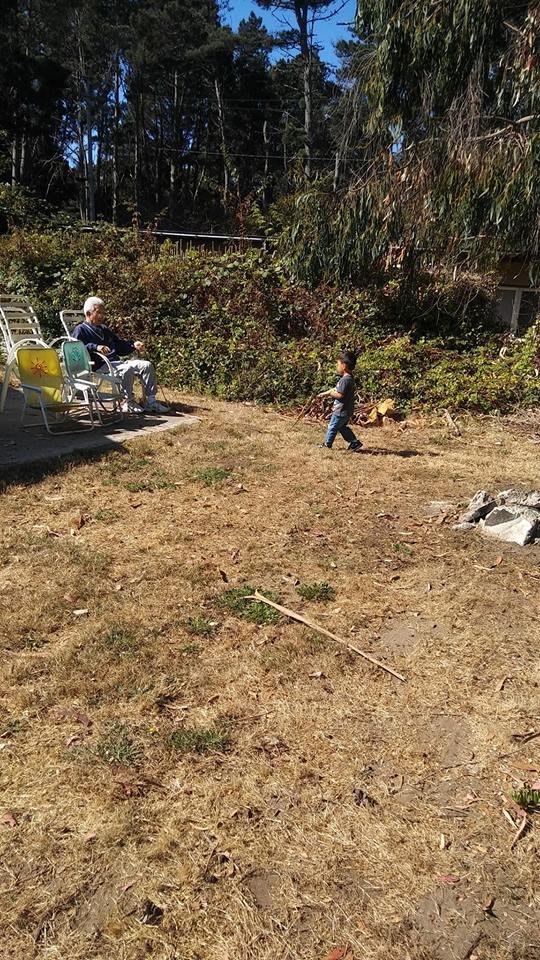 A child playing in a garden while an elderly person relaxes on a sun chair.