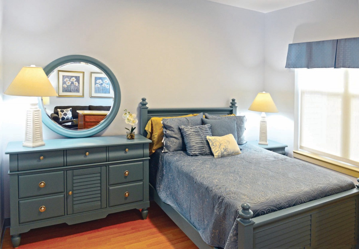 A cozy bedroom with a blue-themed decor, featuring a bed, dresser with mirror, and a lamp.