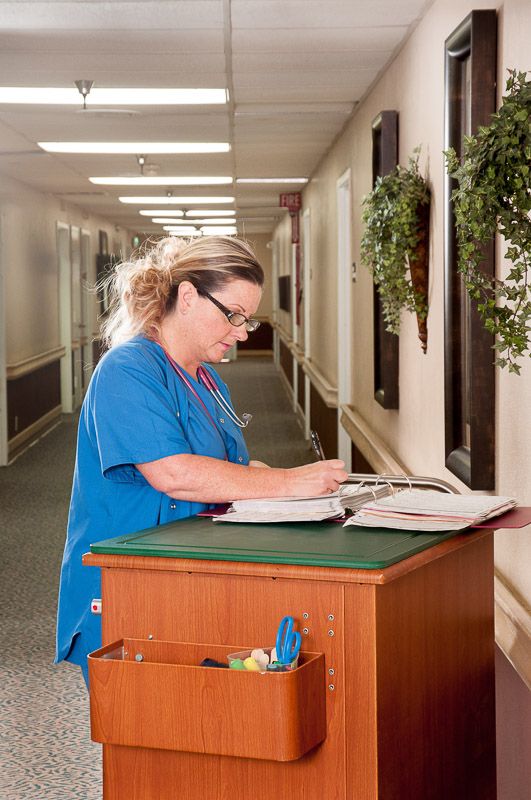 A nurse reviews patient records at a nursing station in a hospital corridor.
