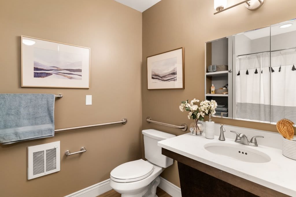 A modern bathroom with neutral tones, featuring a toilet, sink with mirror, and decorative elements like flowers and paintings.