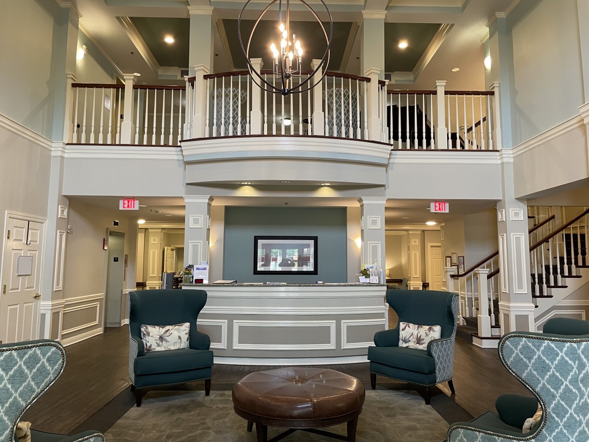 A spacious and elegantly designed lobby featuring a reception desk, upholstered chairs, and a chandelier under a balcony.