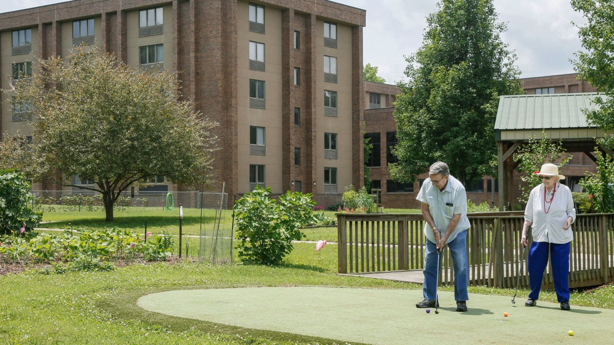 A man playing golf on a small putting green near a residential building.