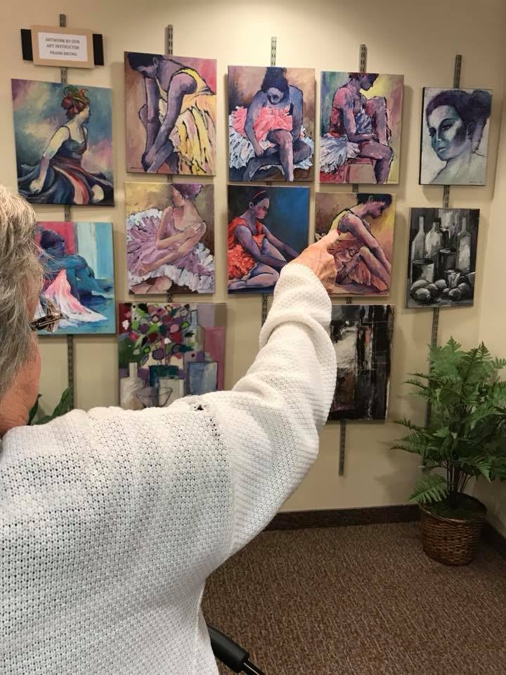A person in a white sweater points towards paintings displayed on a wall in an art gallery.