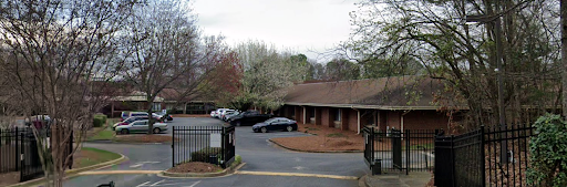 A view of a parking lot near a single-story building, surrounded by trees under a cloudy sky.