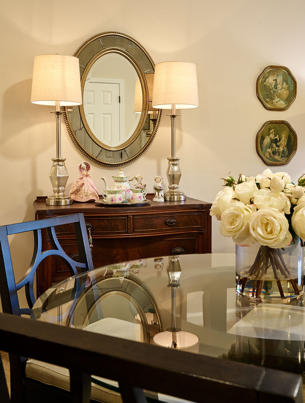 An elegant dining room interior featuring a round glass table, a vase of white roses, and classic decorative elements.