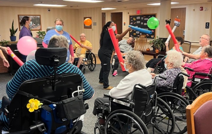 A group of seniors engage in a lively indoor activity with balloons and pool noodles in a nursing home setting.