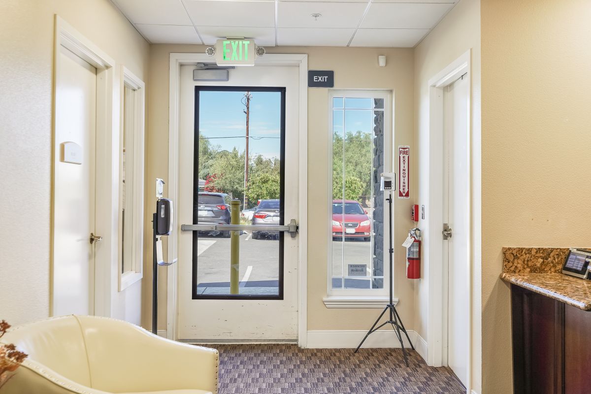 A brightly lit office entrance with a glass door and window, featuring an exit sign and a fire extinguisher.